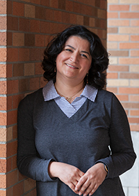 A woman leans against a wall, smiling. It is Dr. Azadeh Yazdan-Shahmorad.